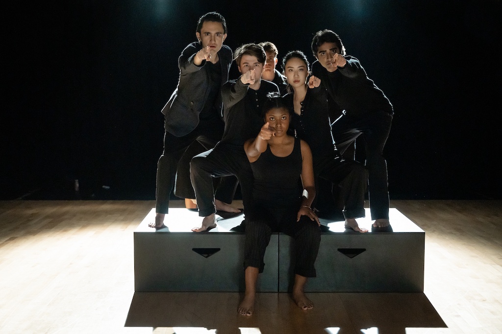 Group of actors stand or sit closely together on two black risers while pointing at the camera during the final presentation of The Winter's Tale.